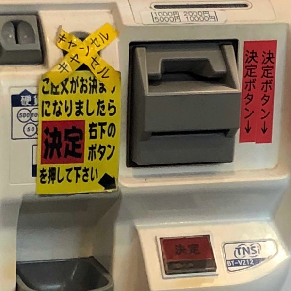 写真: 蒙古タンメン中本高田馬場店の券売機。注意書きが貼られており、その横には「決定ボタン」というテープが2枚貼られている。また、あるボタンの上には「キャンセル」と書かれたテープが交差させて貼られてある。